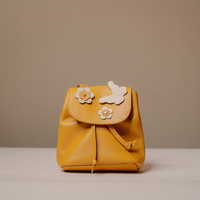 A mustard yellow leather mini backpack with a flap closure, decorated with three white leather flower and butterfly embellishments, set against a neutral beige background.