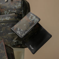 A trifold slender wallet with a camouflage pattern displayed in front of a backpack and a watch. The wallet is predominantly black with camouflage accents.