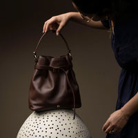 A person holding a brown leather bucket bag with a dark-colored strap, displayed against a neutral background.