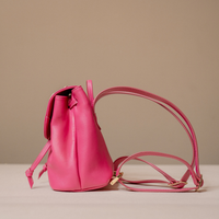 A small bright pink leather bucket bag with a matching shoulder strap and gold-tone hardware, photographed on a neutral beige studio background.