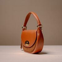 A structured brown leather handbag with a curved top handle, gold-tone hardware, and a flap closure featuring a central gold turn-lock clasp, photographed on a neutral beige studio background.