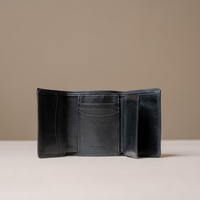 An open black leather bi-fold wallet with multiple card slots and an interior pocket, displayed on a light beige surface against a neutral taupe background.