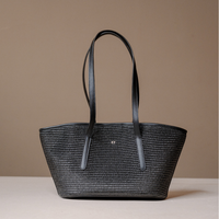 A black woven tote bag with long black handles and a small silver logo at the center, displayed on a light beige surface against a solid taupe background.