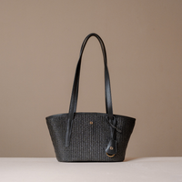 A black woven tote bag with long shoulder straps and a small gold-tone logo charm, displayed on a light beige surface against a solid taupe background.