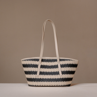 A woven straw tote bag with horizontal black and cream stripes and two long cream shoulder straps, displayed on a light surface against a solid taupe background.