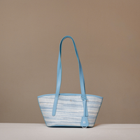 A light blue and white striped tote bag with solid light blue leather handles, standing on a light surface against a solid taupe background.