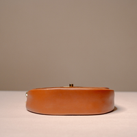 A small oval leather clutch bag in a warm tan or cognac brown, featuring a gold-tone clasp, resting on a light surface against a neutral beige background.