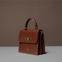 Slightly Side View of Brown Nelda Bag elegant leather handbag with detachable shoulder strap on a gray surface and brown background