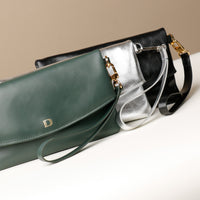 Front view of three leather Karelia Clutch Danilos in green, silver, and black with a visible hand strap and gold accents over a white and beige background
