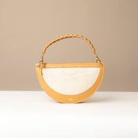 A crescent-shaped shoulder bag with a short handle featuring hand-woven details and a long, adjustable strap. The bag is made with natural fibers and has a zipper closure.
