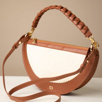 A crescent-shaped shoulder bag with a short handle featuring hand-woven details and a long, adjustable strap. The bag is made with natural fibers and has a zipper closure.
