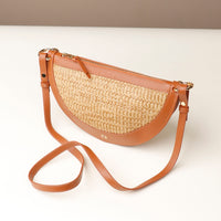 Half-moon shaped handbag with straw texture and brown leather straps on a white background