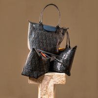 A coordinated set of three handbags in a dark blue and black speckled patterned material, displayed on a stone pedestal against a warm beige background. The set includes a large tote bag with a dark blue leather handle and flap, a medium zippered pouch, and a smaller zippered pouch.