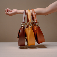 Three small structured leather handbags in brown, tan, and camel hang from a model's extended arm against a warm beige studio background.