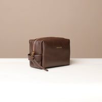 A brown leather toiletry bag with a zipper closure and a small leather strap, displayed against a neutral background.