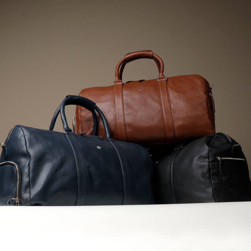 A collection of duffle bags in various colors, featuring a sleek and sophisticated design with sturdy handles and an adjustable leather strap.