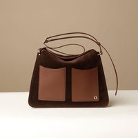 A brown suede shoulder bag with a square shape, featuring a structured design, an edge-cut, fold-over closure with a magnetic clasp, and a brand logo embossed on the bottom right.