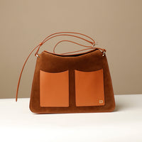 A brown suede shoulder bag with a square shape, featuring a structured design, an edge-cut, fold-over closure with a magnetic clasp, and a brand logo embossed on the bottom right.