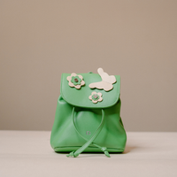 A small mint green leather backpack with a flap closure decorated with two cream flower appliqués and a cream bunny applique, shown on a neutral beige background.