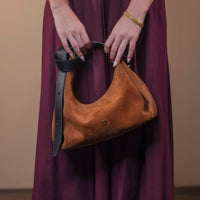 Brown leather handbag held by a person wearing a purple dress against a beige background