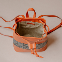 An orange leather bucket backpack with a drawstring closure, featuring a gray patterned band around the middle and an orange leather accent on the drawstring. The interior lining is beige with a subtle geometric pattern.