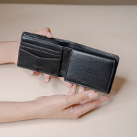 A black leather bifold wallet held open by two hands against a warm beige background. The wallet features multiple card slots on the left and a slip pocket on the right.