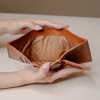 A tan leather pouch or organizer being held open by two hands, showing a patterned brown interior lining on a neutral beige background.
