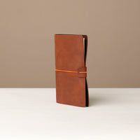 A brown leather journal with a orange elastic band on a white surface.