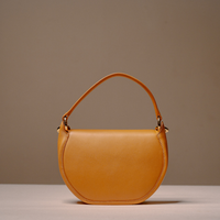 A semi-circular leather handbag in a warm camel tan color, featuring a single top handle and gold-tone hardware, set against a neutral beige background.