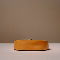 A small oval clutch bag in a warm mustard yellow leather, featuring a top clasp closure, set against a minimalist beige background.