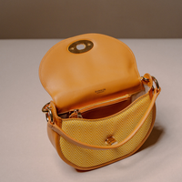 A DKNYOS crescent-shaped shoulder bag in mustard yellow herringbone fabric with tan leather trim and gold-tone hardware, shown open on a neutral beige background.