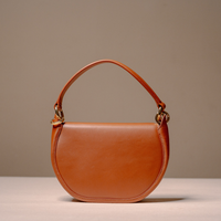 A semi-circular leather shoulder bag in a warm cognac brown, featuring a single top handle and gold-tone hardware, set against a neutral beige background.