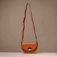 A brown leather crescent-shaped shoulder bag with a gold-tone turn-lock closure and a long adjustable shoulder strap, displayed on a neutral beige background.