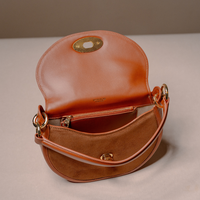 A warm tan leather shoulder bag shown open, displaying its interior zip pocket, suede-like lining, and gold-tone hardware against a neutral beige background.