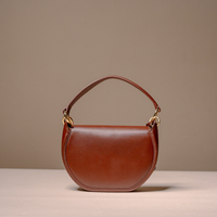 A brown leather crescent-shaped handbag with a curved top handle and gold-tone hardware, displayed on a light beige surface against a neutral taupe background.