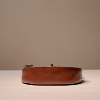 A brown leather belt with gold-tone hardware, shown from the side on a light beige surface against a neutral taupe background.