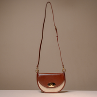 A brown leather crossbody handbag with a curved bottom, a cream-colored textured side panel, and gold-tone hardware, shown against a neutral beige background.