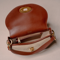 A brown leather shoulder bag shown open, displaying its interior with a zippered pocket, a woven beige lining, and gold-tone hardware against a neutral beige background.