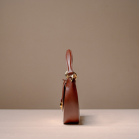 A brown leather handbag shown from the side profile, featuring a structured shape, a top handle, and gold-tone hardware, set against a minimalist beige background.