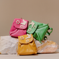 Three small leather backpacks in pink, green, and mustard yellow, each decorated with white flower and butterfly appliqués, arranged on white and tan stones against a warm beige background.