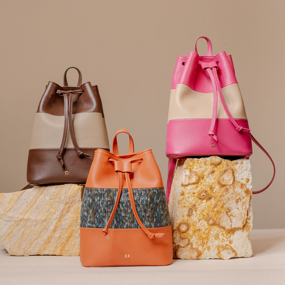 Three leather drawstring backpacks in different color-blocked designs are displayed on stone blocks against a warm beige background. The backpacks feature color blocking in brown and beige, orange and blue-gray, and bright pink and cream.