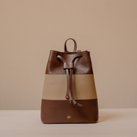 A color-block leather drawstring backpack in dark brown, tan, and beige, featuring a small gold monogram 'D' on the front, set against a neutral beige background.