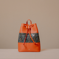 An orange leather drawstring backpack with a wide, patterned blue and brown woven band across the middle, displayed on a neutral beige background.