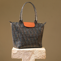 A structured tote bag with a dark blue and brown speckled pattern, featuring a contrasting orange flap and matching orange handle accents, displayed on a stone pedestal against a solid beige background.