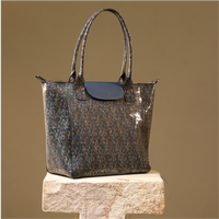 A structured tote bag with a glossy, speckled multicolor finish in shades of blue, brown, and gray, featuring a dark navy flap and handles, displayed on a stone pedestal against a solid tan background.