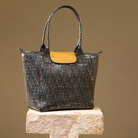 A textured tote bag with a dark blue and brown speckled pattern and a contrasting mustard yellow flap, displayed on a stone pedestal against a solid beige background.