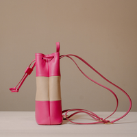 A small leather bucket bag in bright pink and beige color blocking, featuring a matching pink drawstring strap and a long thin shoulder strap, set against a neutral beige background.
