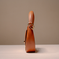 A side profile view of a warm brown leather handbag with gold-tone hardware, standing on a light surface against a neutral beige background.