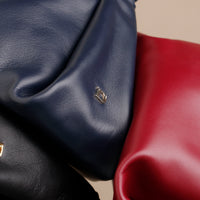 Close-up of three leather bags in navy, black, and red on a beige background.