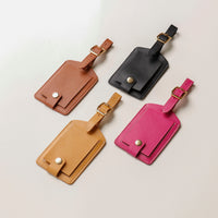 Four leather luggage tags in brown, black, beige, and pink on a light gray background.
